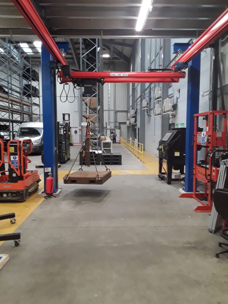Angled underside view of red overhead light crane rails and trolley hoist units fitted beneath a warehouse's white steel roof structure