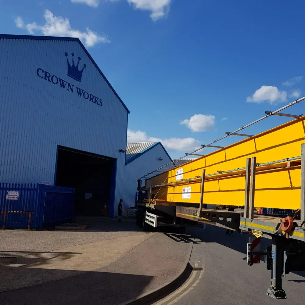 A yellow bridge crane with a central hoist unit, mounted on a steel runway system inside a bright, spacious industrial facility with warehouse racking in the background.