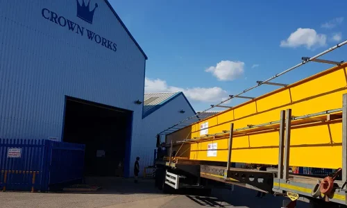 A yellow bridge crane with a central hoist unit, mounted on a steel runway system inside a bright, spacious industrial facility with warehouse racking in the background.